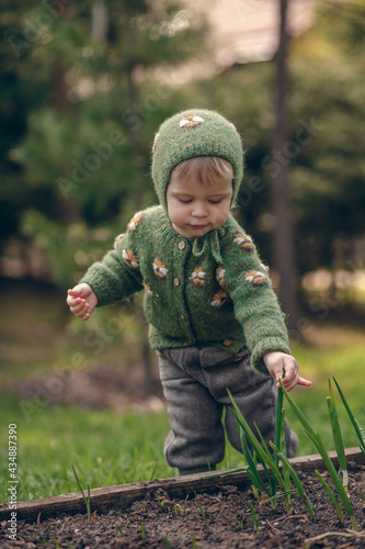 Cute baby girl in green knitted bonnet and sweater is eating green onion from the garden bed. Image with selective focus and toning.