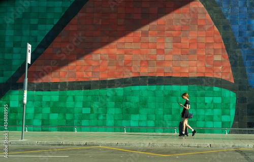 chica joven de pelo corto y rojo camiando con la maleta por el aeropuerto de Barcelona hacia la parada del autobus mirando el movil con un mosaico gigante detras  photo