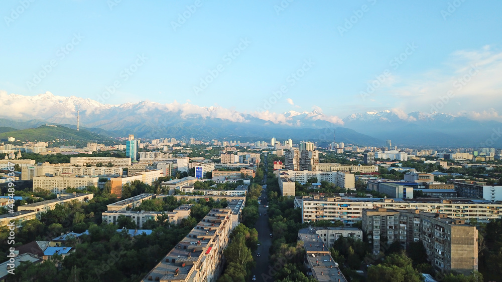 Bright color sunset over the city of Almaty. Huge clouds over the mountains and the city shimmer from bright blue to yellow and dark blue. Tall houses and green trees, cars driving on the roads.