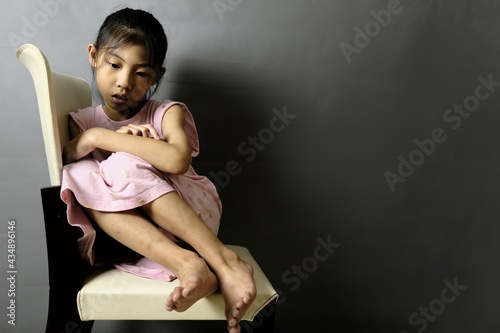 A sad abused girl sitting on a chair in a dark room, feeling lonely and depressed with dark gray shadow in the background.
