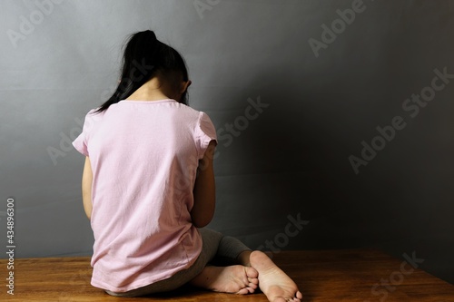 Child abuse concept. The back view of an orphanage girl sitting alone in dark room with her face to a gray dark wall, feeling lonely and scare, crying.
