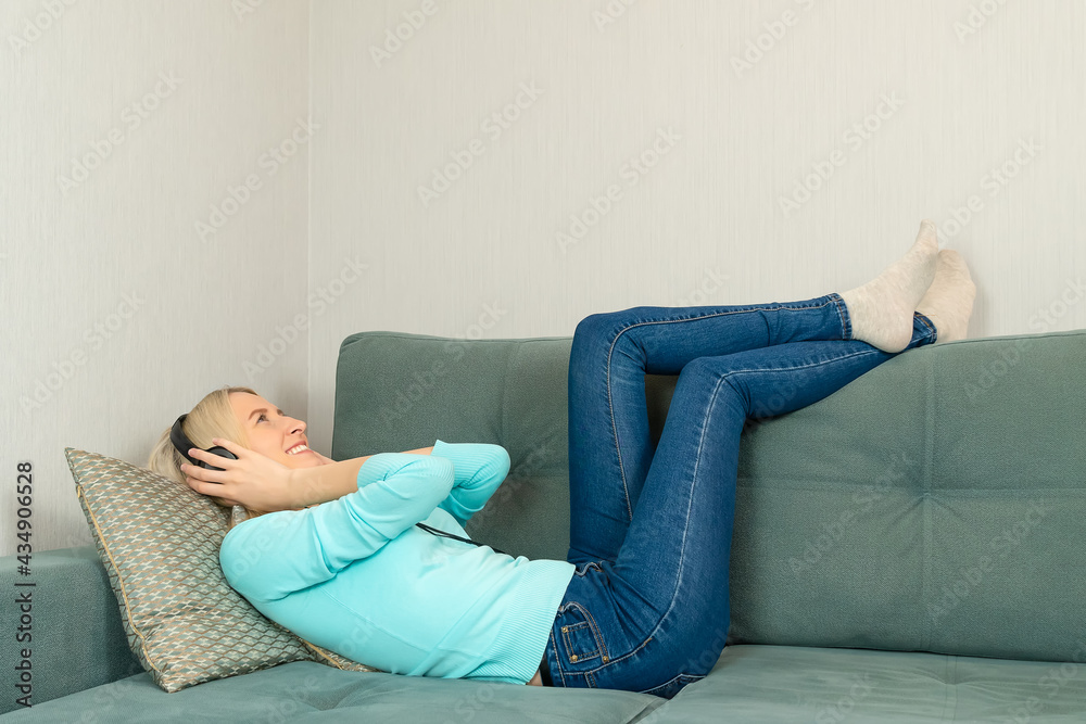 Young blonde woman in bright clothes is lying on the sofa with large headphones listening to music and smiling