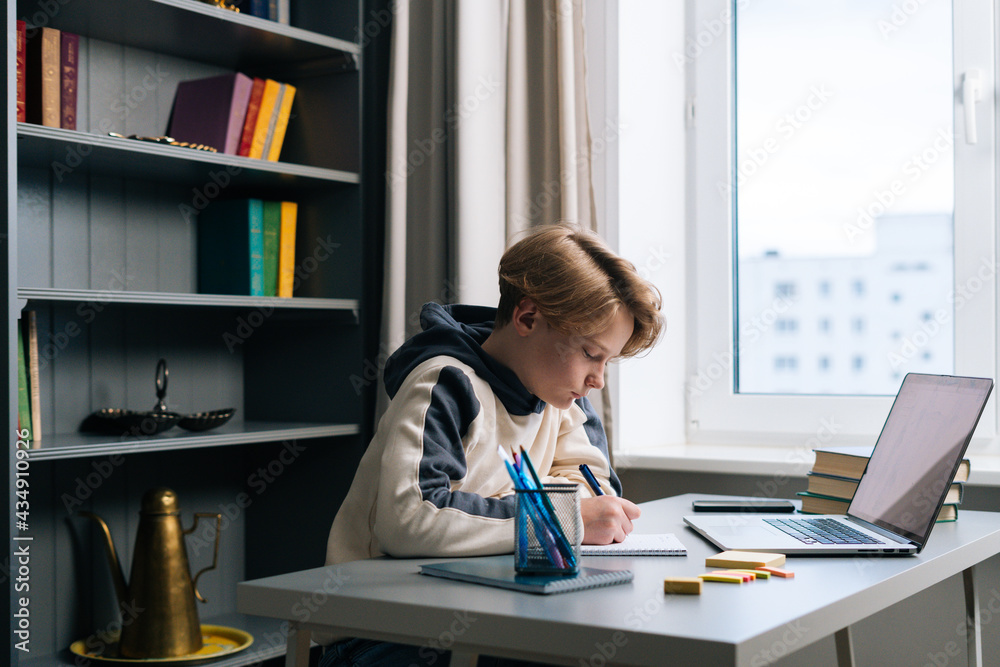 Side view of pupil boy making notes in copybook with pen sitting at desk with laptop computer via distance online class. Child schoolboy doing homework at home new window. Concept of remote education. Side view of pupil boy making notes in copybook with pen sitting at desk with laptop computer via distance online class. Child schoolboy doing homework at home new window. Concept of remote education.