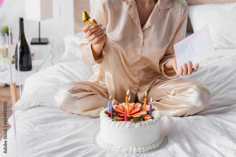 Cropped view of woman in pajama holding party horn and greeting card near birthday cake on bed Cropped view of woman in pajama holding party horn and greeting card near birthday cake on bed
