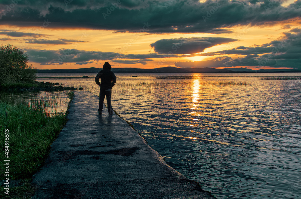Beautiful orange sunset scenery with man standing on the dock at Corrib lake in Galway, Ireland 