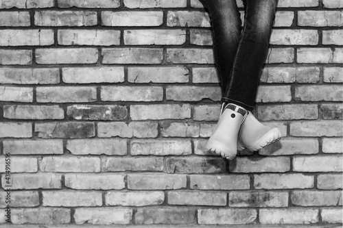 crossed legs of a young girl in jeans and orange galoshes sitting on a brick wall against a brick wall background with copy space, black and white photo © Andrey Solovev