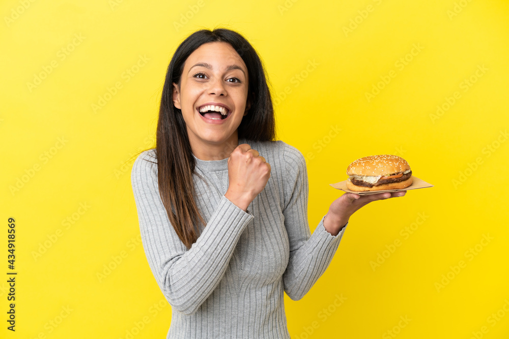 Young caucasian woman holding a burger isolated on yellow background celebrating a victory Young caucasian woman holding a burger isolated on yellow background celebrating a victory