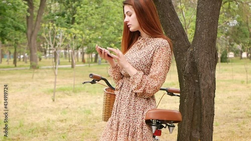A beautiful woman uses her phone to communicate with friends, texting, social media and internet. Walk through the park in the city with a bike for hire. Rest in nature, active lifestyle. general view photo