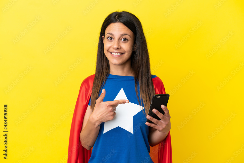 Super Hero woman isolated on yellow background using mobile phone and pointing it Super Hero woman isolated on yellow background using mobile phone and pointing it