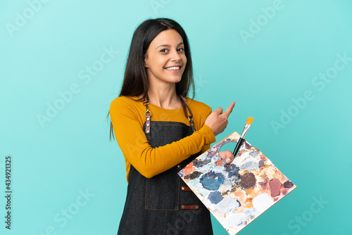 Young artist caucasian woman holding a palette isolated on blue background pointing back photo