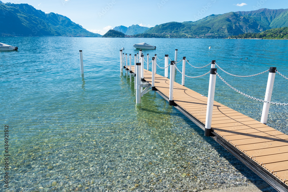 A diagonal gangway leading to a small touristic pier with a boat on the turquoise waters of Lake Como. Menaggio, Italy. At the opposite coast of the lake, the cape of Bellagio is visible. Copy-space.