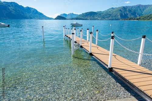 A diagonal gangway leading to a small touristic pier with a boat on the turquoise waters of Lake Como. Menaggio, Italy. At the opposite coast of the lake, the cape of Bellagio is visible. Copy-space.