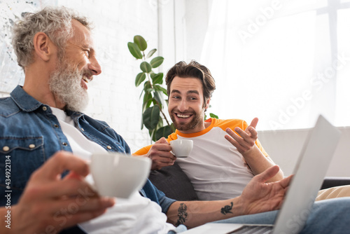 Smiling man with coffee pointing at blurred laptop near father at home