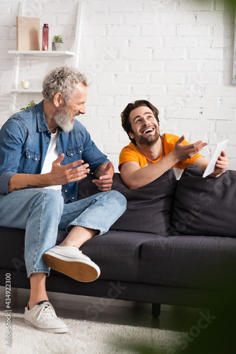 Smiling man pointing at digital tablet near middle aged father on couch