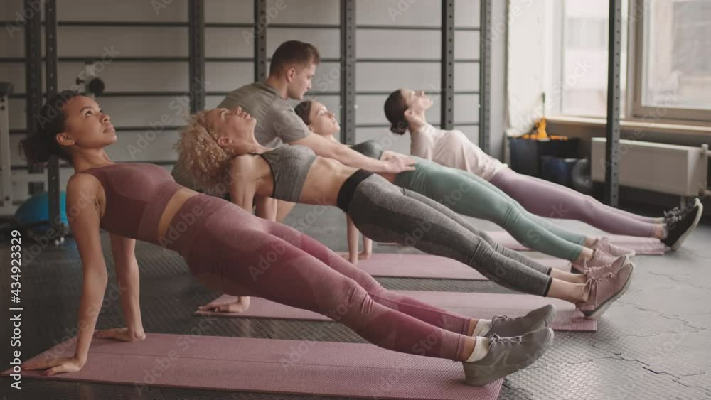 Full shot of four young Mixed-Race and Caucasian women doing reverse plank on yoga mats in gym at daytime, male coach helping them with technique