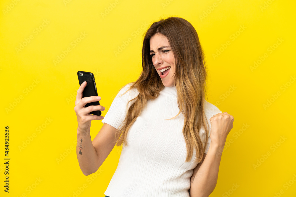 Caucasian woman isolated on yellow background using mobile phone and doing victory gesture