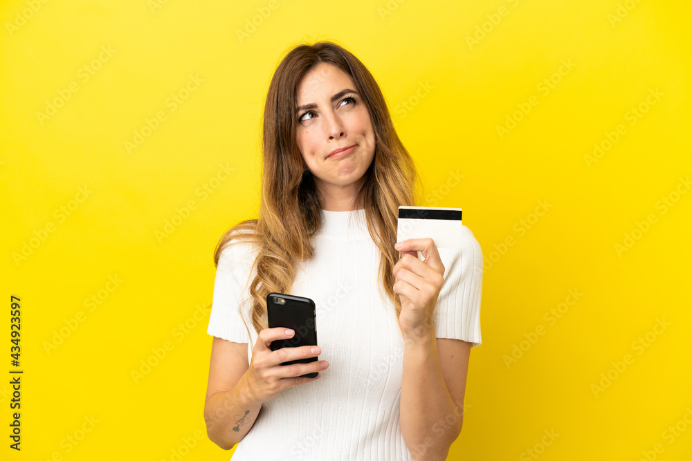 Caucasian woman isolated on yellow background buying with the mobile with a credit card while thinking Caucasian woman isolated on yellow background buying with the mobile with a credit card while thinking