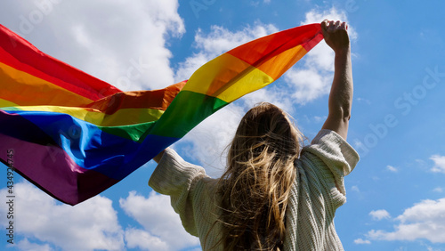 Blonde, lesbian, woman holding a rainbow LGBT gender identity flag on sky background with clouds on a sunny day and celebrating a gay parade