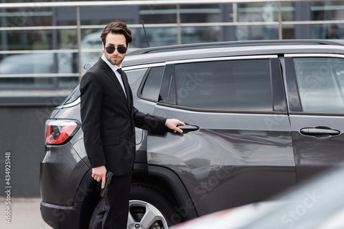 safeguard in sunglasses and suit holding gun near modern auto