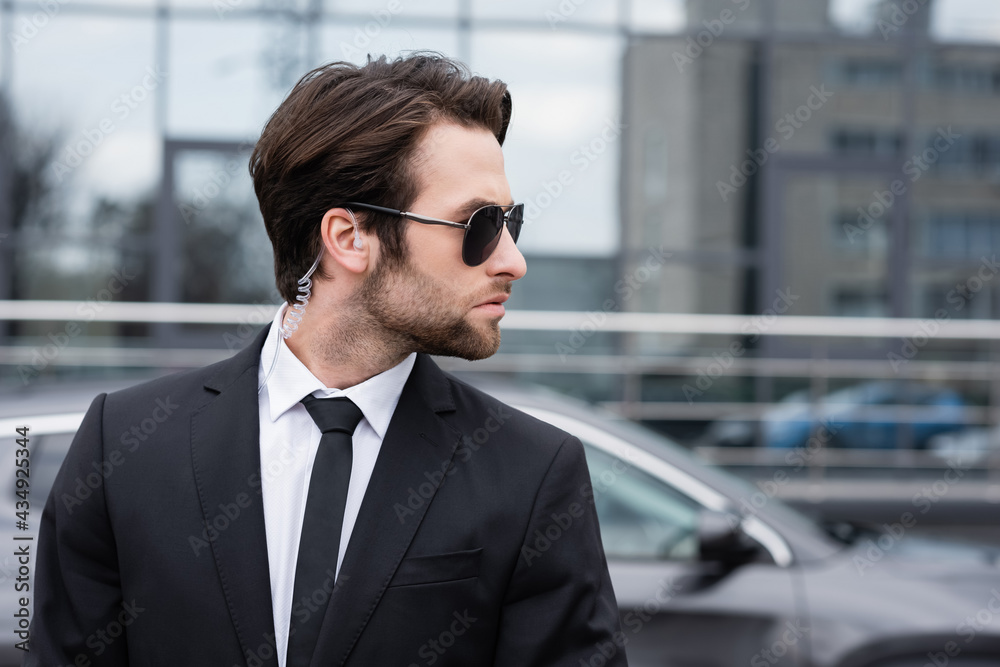 side view of bearded bodyguard in suit and sunglasses with security earpiece near modern car side view of bearded bodyguard in suit and sunglasses with security earpiece near modern car