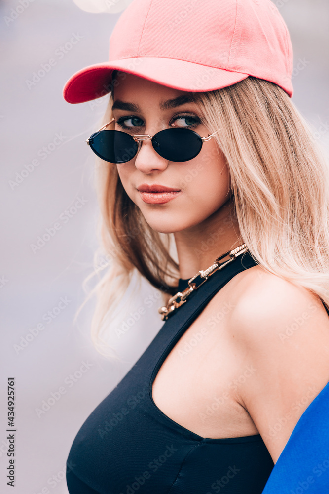 Young sexy blonde hipster woman posing on the street. Wearing blue stylish jacket, jeans and baseball hat and sunglasses. Young sexy blonde hipster woman posing on the street. Wearing blue stylish jacket, jeans and baseball hat and sunglasses.