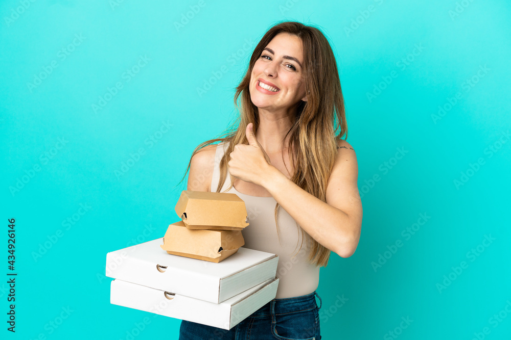 Caucasian woman holding pizzas and burger isolated on blue background giving a thumbs up gesture Caucasian woman holding pizzas and burger isolated on blue background giving a thumbs up gesture