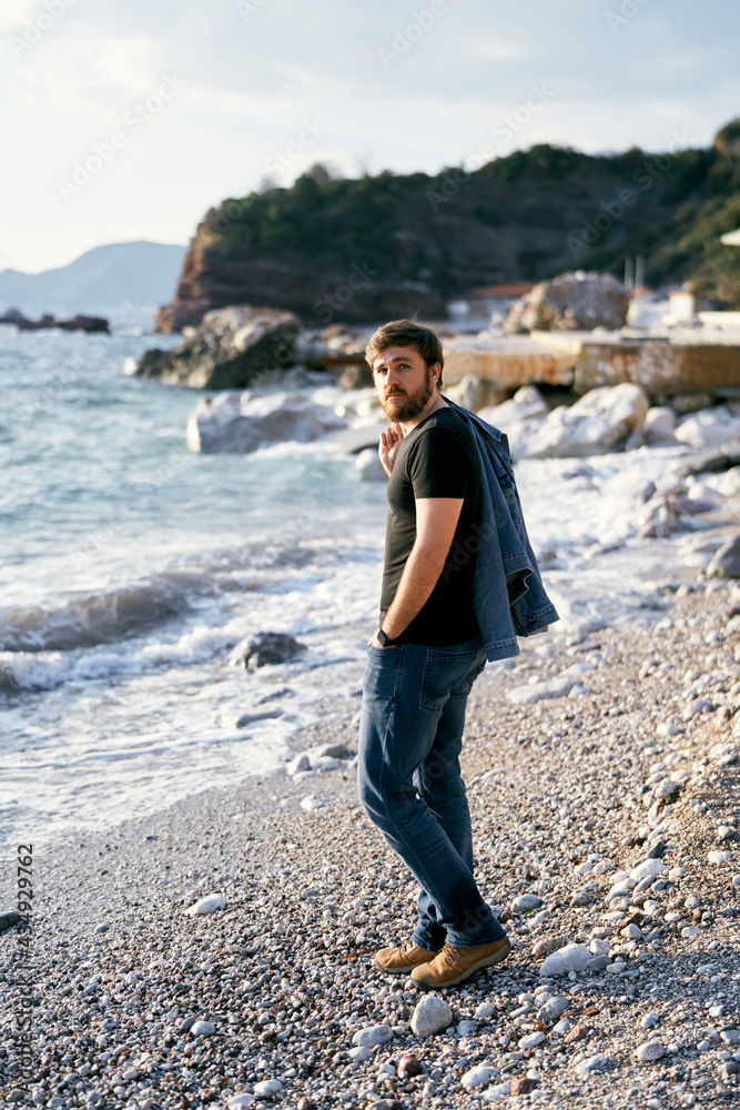Pensive man stands on a pebble beach with a denim jacket over his shoulder against the background of the sea and rocks