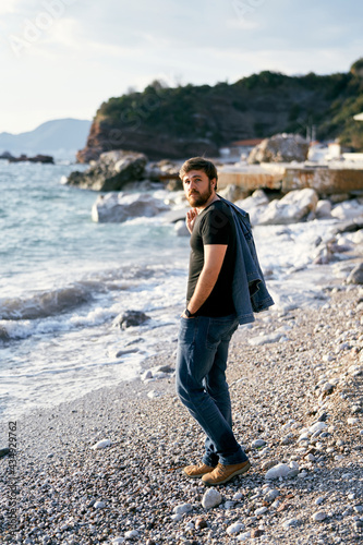 Pensive man stands on a pebble beach with a denim jacket over his shoulder against the background of the sea and rocks © Nadtochiy