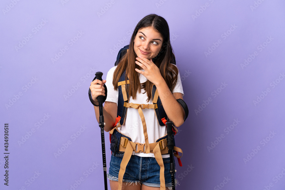 Young woman with backpack and trekking poles isolated on purple background looking up while smiling Young woman with backpack and trekking poles isolated on purple background looking up while smiling