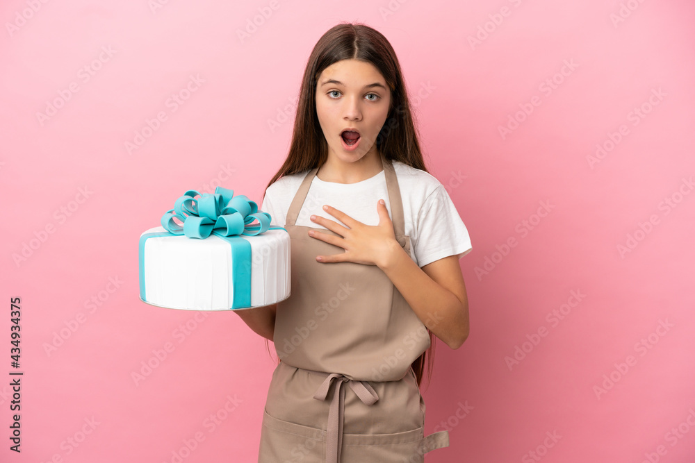 Little girl with a big cake over isolated pink background surprised and shocked while looking right