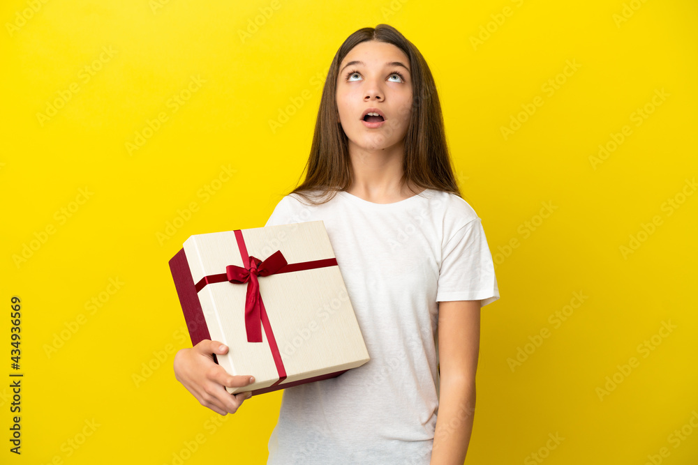 Little girl holding a gift over isolated yellow background looking up and with surprised expression Little girl holding a gift over isolated yellow background looking up and with surprised expression