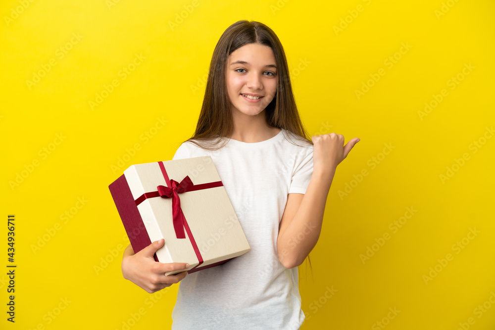 Little girl holding a gift over isolated yellow background pointing to the side to present a product Little girl holding a gift over isolated yellow background pointing to the side to present a product