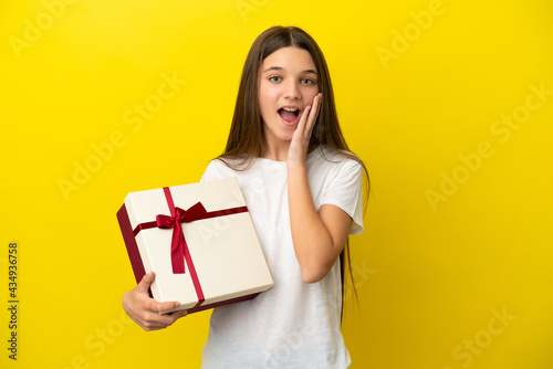 Little girl holding a gift over isolated yellow background with surprise and shocked facial expression