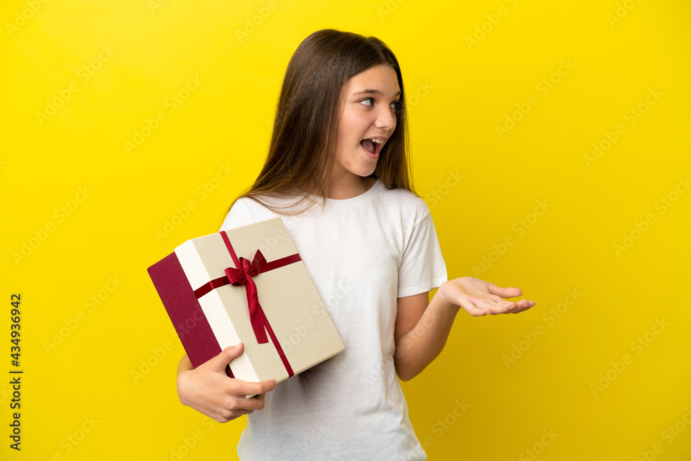 Little girl holding a gift over isolated yellow background with surprise expression while looking side Little girl holding a gift over isolated yellow background with surprise expression while looking side