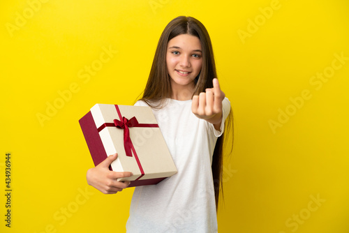 Little girl holding a gift over isolated yellow background doing coming gesture