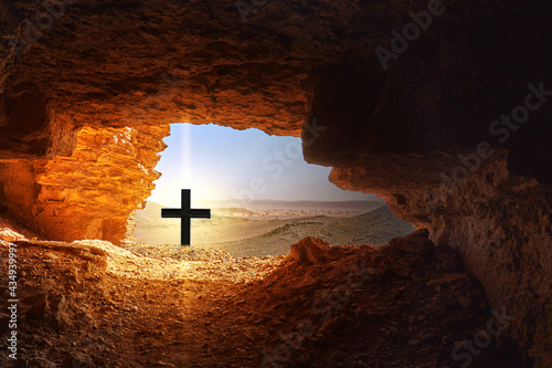 Black cross with bright light in front of dark cave in the dry hostile desert with boulders