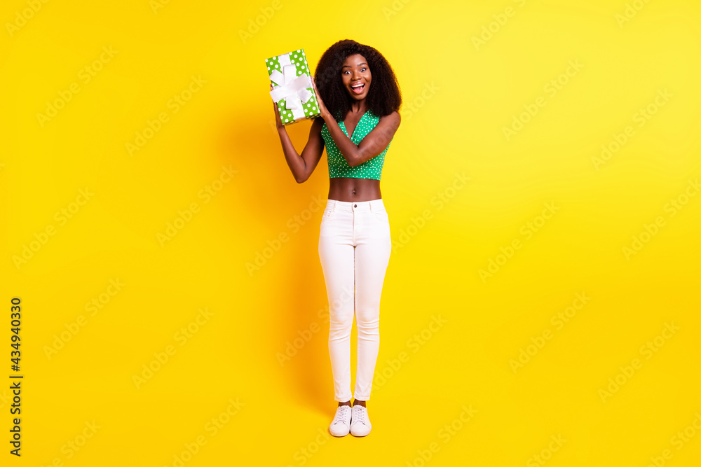 Full length body size photo funky girl smiling trying to guess what inside gift box isolated vivid yellow color background Full length body size photo funky girl smiling trying to guess what inside gift box isolated vivid yellow color background