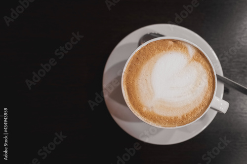 a white cappuccino coffee cup with a heart - shaped pattern on the foam. a cup and saucer on a dark wooden table in a cafe or restaurant