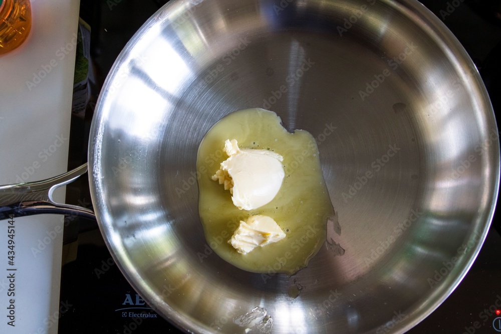 A portrait of two pieces of butter lying in a stainless steel frying pan on an induction cooker. The dairy product is melting so the cook can fry some meat or prepare some other type of food in it.