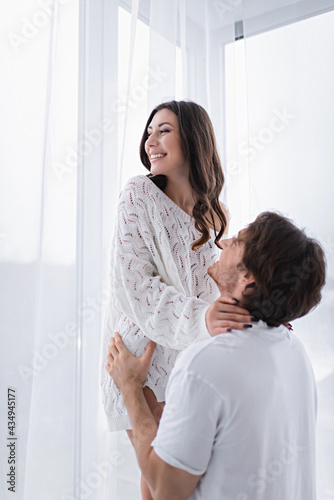 Smiling woman embracing blurred boyfriend near window