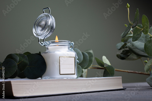 Glass candle jar with metal lid and white label. Burning candle. Nearby eucalyptus branches and gray concrete background 