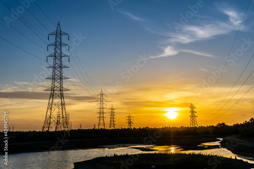 Silhouette effect of high voltage electric towers with electricity transmission power lines, located near riverside and sunset sky background