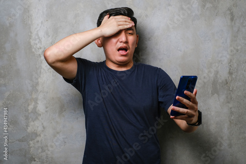 Young Japanese man using smartphone stressed with hand on head, shocked with shame and surprise face. Fear and upset for mistake.