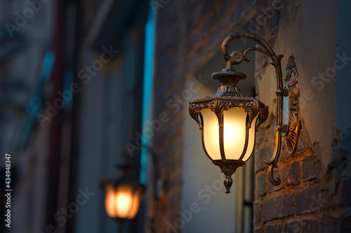 Lantern with yellow warm light on facade of building. Twilight on city street, building illumination. Retro lantern lighting, warm light glow. Street lights, illumination and vintage lantern