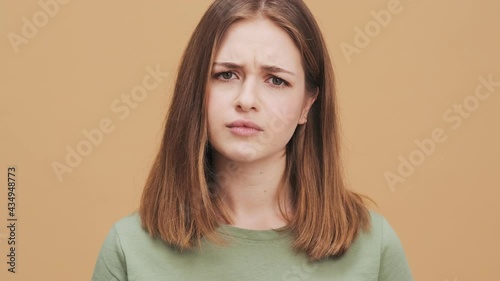 A serious young woman is shaking her head negatively while listening the viewer standing isolated over a beige wall in the studio
