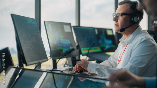 Male Air Traffic Controller with Headset Using Touchscreen Table Panel in Airport Tower. Office Room is Full of Computers with Navigation Screens, Airplane Departure and Arrival Data for the Team. photo