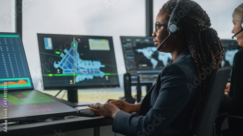 Female Air Traffic Controller with Headset Talk on a Call in Airport Tower. Office Room is Full of Desktop Computer Displays with Navigation Screens, Airplane Departure and Arrival Data for the Team.