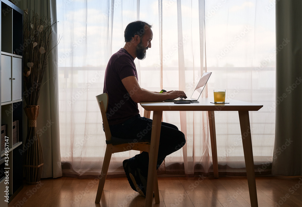 backlit portrait of middle-aged bearded man working with his laptop by the window of his home backlit portrait of middle-aged bearded man working with his laptop by the window of his home