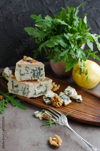 Gorgonzola on a wooden plate next to arugula and walnuts. Cheese with a noble blue mold on a gray table and
dark background.