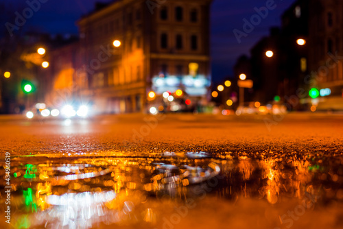 Nights lights of the big city, the night intersection between the houses on which cars travel. Close up view of a puddle on the level of the hatch